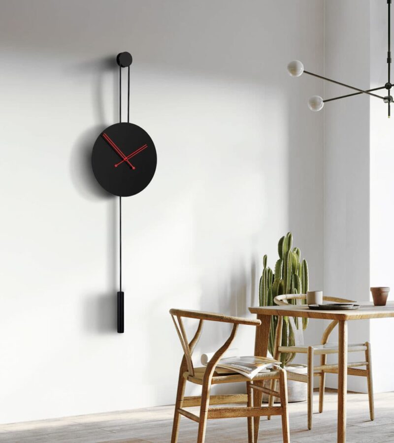 Duvar Saati modelleri arasında olan kırmızı akrep yelkovanlı siyah Halatlı siyah dekoratif saat Avvolo 45 sandalye ve masa olan odada duvarda asılı durmakta